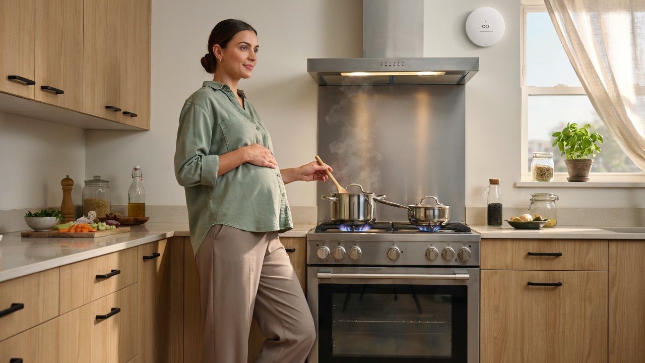 pregnant woman standing in a well-ventilated kitchen with a gas stove, range hood running, and a carbon monoxide detector visible on the wall, illustrating gas appliance safety during pregnancy